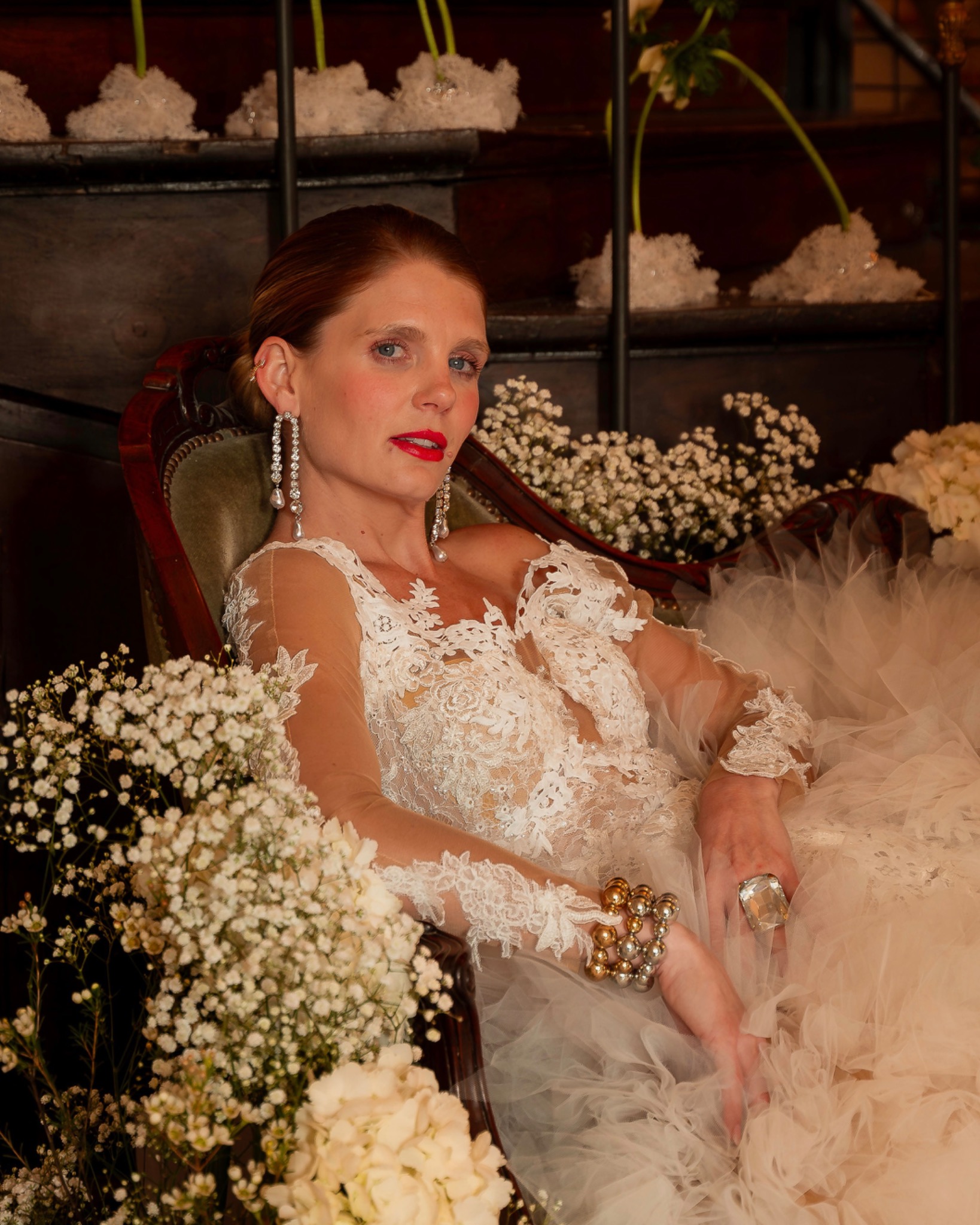 Bride seated among white flowers in a lace gown, with fresh skin, softly flushed cheeks and a bright red lip.