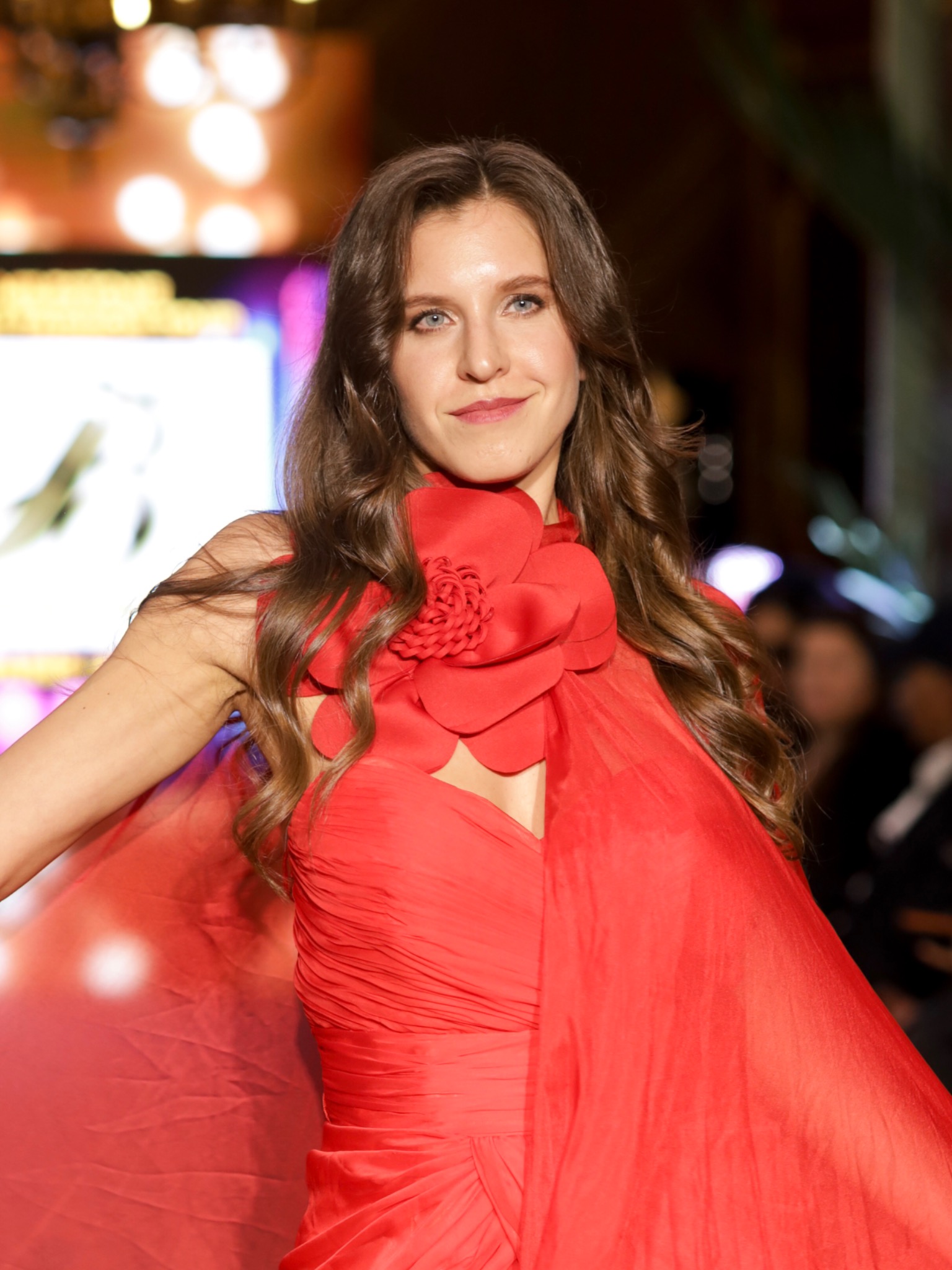Runway portrait with warm glowing skin, soft brown eye makeup and a rosy lip, in a red draped gown.