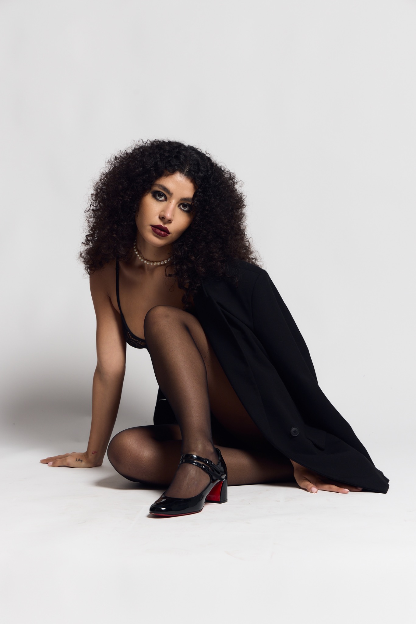 Full-length studio portrait with smoked eyes and deep burgundy lips, seated in a black tailored jacket on a pale backdrop.