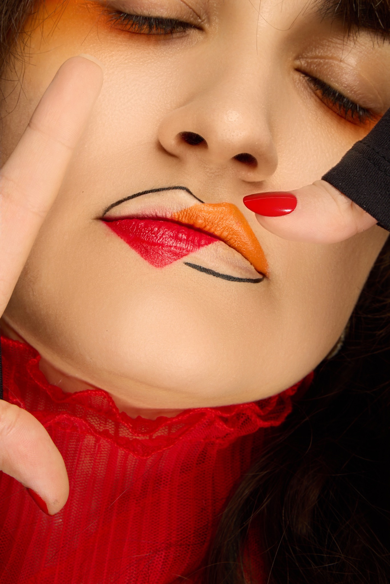 Editorial close-up of a graphic two-tone red and orange lip outlined in black liner, with warm shadow on the eyes.