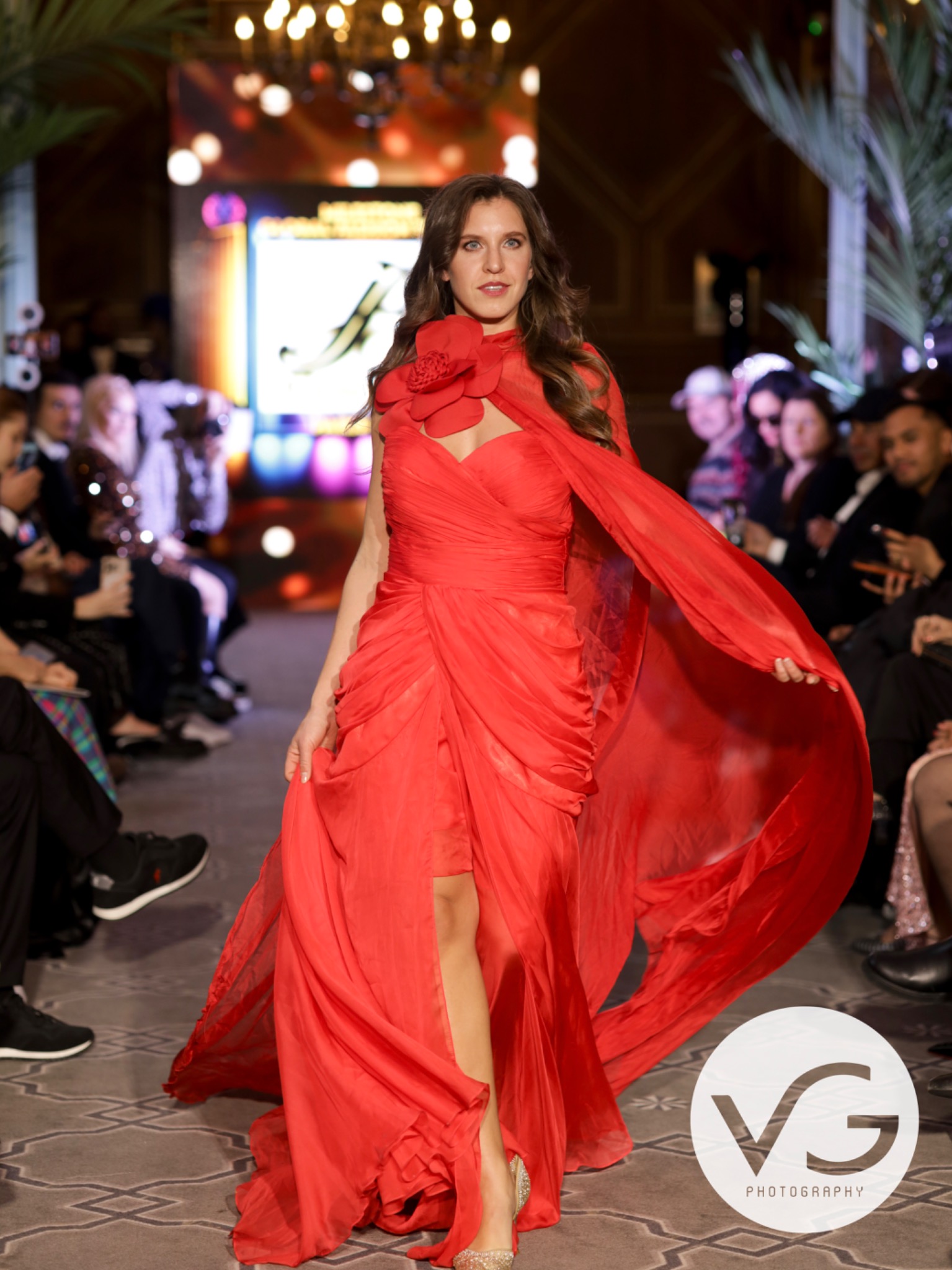 Full-length runway shot with soft luminous makeup and defined eyes, in a flowing red gown with a floral neckline.