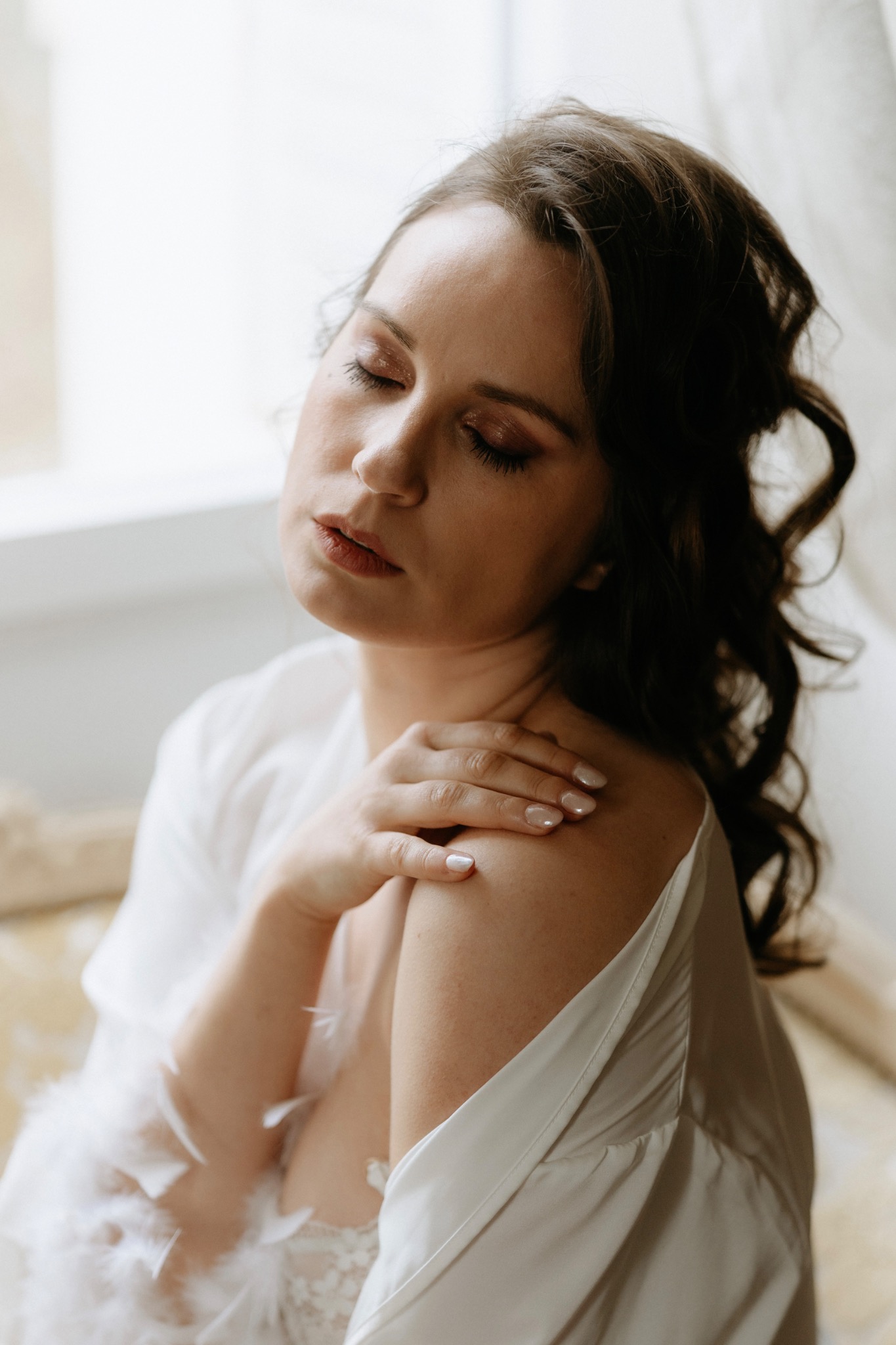 Bride in a white silk robe by a bright window, with soft shimmering eye makeup, glowing skin and a muted red lip.