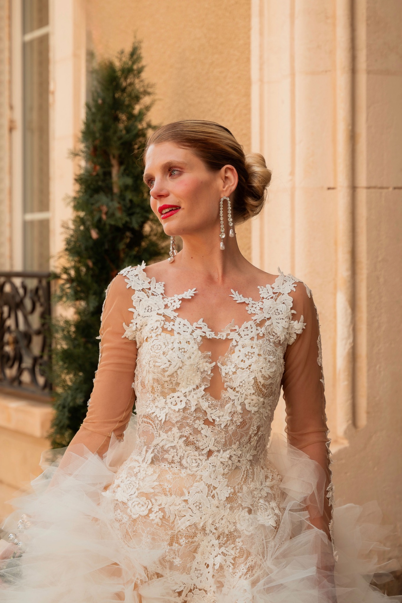 Bride outdoors in warm daylight wearing a lace gown, with a sleek low bun, luminous skin and a vivid red lip.
