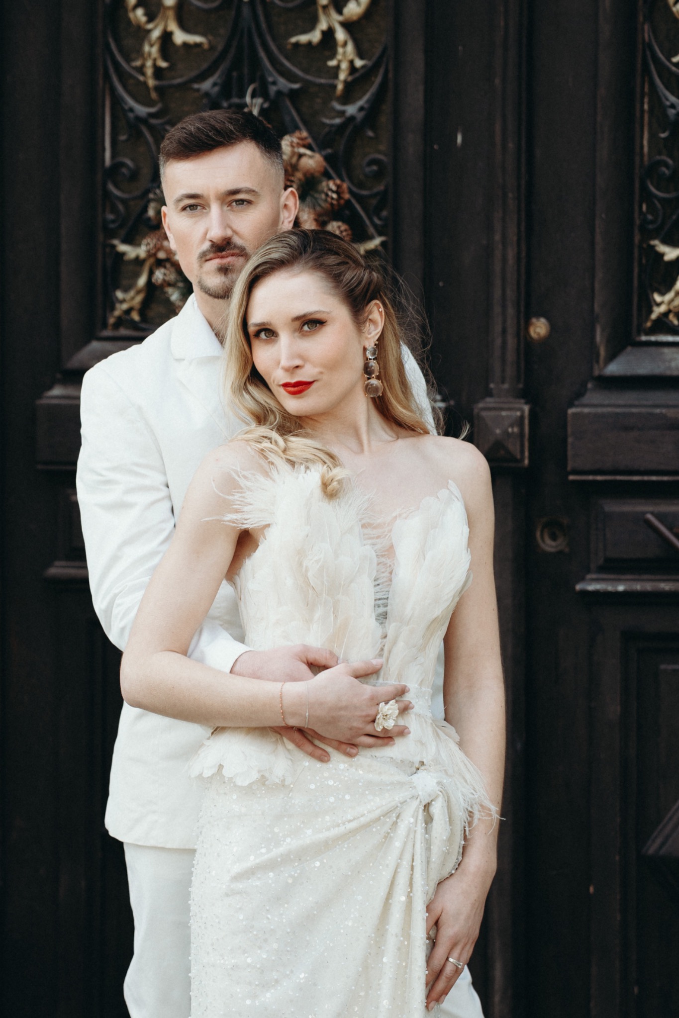 Couple standing before a dark carved door, the bride in a feathered gown with softly defined eyes and a red lip.