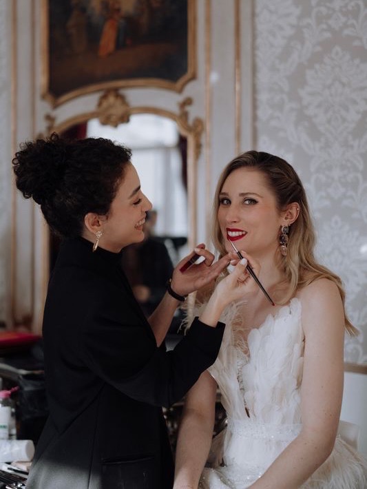 Behind the scenes: lipstick being applied with a brush to a smiling bride in a gilded room.
